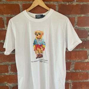 Vit Polo Bear t-shirt från Ralph Lauren - Vit t-shirt från Polo Ralph Lauren med ikoniskt Polo Bear-tryck på bröstet. Klassisk rund hals och kortatarmad. en skön känsla och snygg passform. Perfekt statement-plagg för dig som gillar en snygg T-shirt 