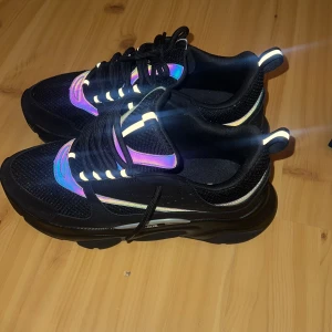 Svarta Multifärgade Dior B22 skor - Säljer ett par svarta Dior sneakers med chunky sula och reflekterande detaljer i lila, blått och grönt. Skorna har snörning, meshpartier och coola holografiska inslag som syns i ljuset. Kommer med original dustbag från Dior. Priset är ej hugget i sten så ställ hur många frågor ni vill!☺️
