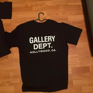 Svart Gallery Dept. t-shirt - Svart t-shirt från Gallery Dept. med vit text på bröstet och stor logga på ryggen. Klassisk passform med rund hals och korta ärmar. Tillverkad i mjuk bomull för en skön känsla. Perfekt för dig som gillar streetwear och tydliga loggor.