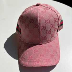 Rosa keps - Säljer en rosa keps med klassiskt GG-monogram över hela kepsen. Kepsen har gröna och röda ränder på sidorna och justerbar rem bak. Tillverkad i bomull och polyester, perfekt för dig som vill sticka ut med en lyxig accessoar.