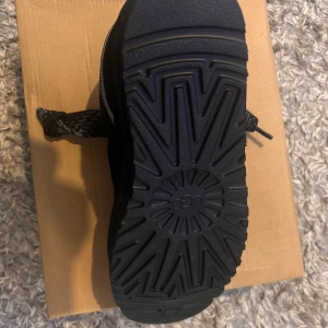 UGGS LOWMEK SVART - Säljer ett par svarta UGG sneakers med grov, mönstrad sula och breda snören i svart och grått mönster. Skorna har detaljer i mocka och mesh, samt en reflexdetalj på sidan. UGG-logga på plösen och sidan. Perfekta för dig som gillar chunky streetstyle.