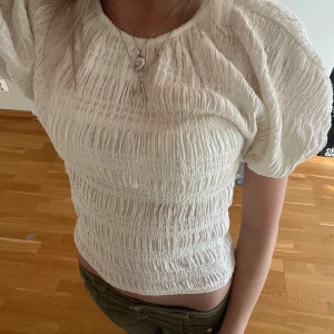 Vit smockad blus med puffärm - Supersöt vit blus med smockad struktur och puffiga korta ärmar. Toppen är croppad och har en rund halsringning. Perfekt för dig som gillar en feminin och trendig stil. Materialet känns lätt och luftigt.