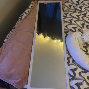 Spegel  - Detta är en helkroppsspegel med vit ram. Spegeln är i gott skick men har en mindre spricka i ett av hörnen på ramen, vilket syns på närbilden. Perfekt för sovrum eller hall. Ger ett modernt och stilrent intryck.