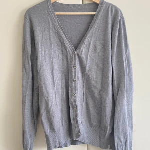 Grå kofta med knappar - En enkel och stilren grå kofta med v-ringning och knäppning framtill. Koftan har långa ärmar och ribbade muddar vid ärmslut och nederkant. Perfekt att slänga över en t-shirt eller linne för en chill look.