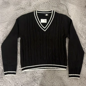 Svart kabelstickad tröja H&M  - Svart kabelstickad tröja från H&M med vita ränder på v-ringad krage, ärmslut och nederkant. Långärmad och med klassisk preppy look, perfekt till jeans eller kjol. Mjuk och skön känsla, passar dig som gillar stilrena detaljer.