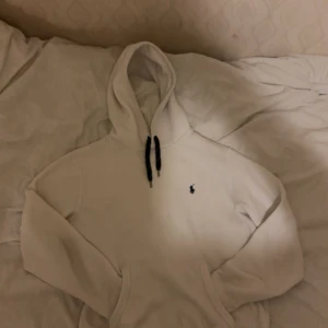 Vit hoodie från Polo Ralph Lauren - Vit hoodie från Polo Ralph Lauren med svart broderad logga på bröstet och svarta snören i huvan. Klassisk känguruficka framtill och mjukt material som känns skönt mot huden. Perfekt för dig som gillar stilrena och tidlösa plagg.