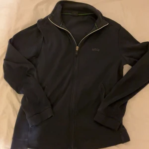 Svart ziptröja från BOSS - Svart långärmad ziptröja från BOSS med diskret logga på bröstet och hel dragkedja framtill. Tröjan har ståkrage och är tillverkad i mjuk bomullsmix, perfekt för lager-på-lager. Snygga detaljer vid kragen och klassisk passform.