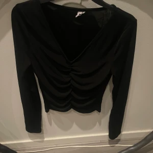 Svart långärmad topp från Nelly - Svart långärmad topp från Nelly med v-ringning och snygg rynkad detalj framtill. Toppens material är stretchigt och tunt, vilket ger en smickrande passform. Perfekt att styla med jeans eller kjol för en trendig look.