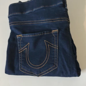 Mörkblå True Religion jeans - Säljer ett par mörkblå jeans från True Religion med ikoniska bakfickor och kontrastsömmar. Klassisk femficksmodell med raka ben och snygg passform. Perfekta för dig som gillar streetwear och vill sticka ut med detaljer.