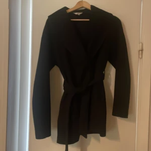 Svart omlottklänning från ONLY - Svart jacka från ONLY med bälte i midjan. Kappan har en klassisk krage och är i en stilren, enkel design som passar perfekt för dig som gillar minimalistiskt mode.