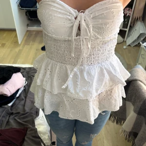 Vit volangig bandeau tubtopp - Köpt i Spanien på marknad så vet ej märket 🥰Supersöt vit bandeau tubtopp med broderade blommor och volanger. Toppen har smockad midja, knytning framtill och är helt axelbandslös. Perfekt för sommarens varma dagar och ger en romantisk vibe. 