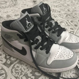 Nike Air Jordan 1 Mid grå/vit - Säljer ett par Nike Air Jordan 1 Mid i snygg grå och vit färg med svarta detaljer och snörning. Skorna har klassisk Jordan-logga på sidan och tungan, samt svart swoosh. Tillverkade i skinn och syntet, med perforerad tå och platt sula.