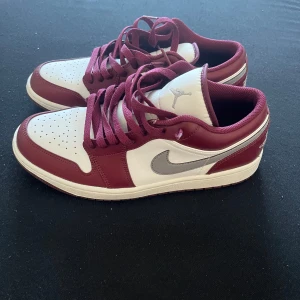Nike Air Jordan 1 Low Bordeaux/Vit - Säljer ett par Nike Air Jordan 1 Low i bordeauxrött och vitt med silverfärgad swoosh. Skorna har klassisk rund tå, perforerad tåbox och snörning. Ovandelen är i skinn och de har en platt sula. Perfekta för dig som vill sticka ut med stilrena sneakers.