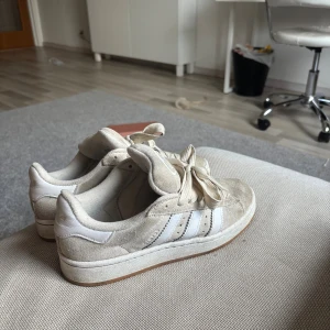 Beige Adidas Campus sneakers mocka - Säljer ett par beige Adidas Campus sneakers i mocka med klassiska vita ränder och gummisula. Skorna har snörning och rund tå, samt Adidas logga på plösen. Perfekta för dig som gillar en clean och stilren look med retrovibbar.