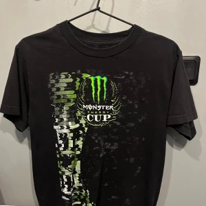 Monster Energy svart t-shirt - Svart t-shirt med coolt Monster Energy Cup-tryck i grönt, vitt och svart på framsidan. Klassisk rund hals och korta ärmar. Materialet känns som bomull och passformen är normal. Perfekt för dig som gillar motorsport eller streetwear. Storlek S 