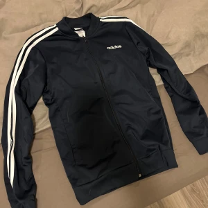 Svart Adidas bombertröja med vita ränder - Snygg svart bomberjacka från Adidas med klassiska vita ränder längs ärmarna och dragkedja framtill. Jackan har ribbade muddar och står för en sportig och clean stil. Perfekt för dig som gillar streetwear och sportmode.
