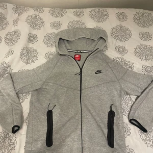 Grå Nike hoodie med dragkedja - Säljer en grå Nike hoodie med hel dragkedja och svarta detaljer. Tröjan har huva, två fickor med dragkedja och långärmade ärmar. Perfekt för chill eller träning, klassisk Nike-logga på bröstet och mjukt material.