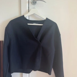 Tunnkofta/kappa från Gina Tricot - Säljer en svart omlottkofta från Gina Tricot med v-ringad hals och lång ärm. Koftan är croppad och har en stilren, enkel design som passar perfekt till jeans eller kjol. Materialet är mjukt och stickat för en skön känsla.