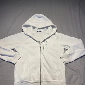 Vit hoodie från Sail Racing med dragkedja - Vit hoodie från Sail Racing med broderad logga på bröstet och stor text på ryggen. Tröjan har huva, hel dragkedja och två fickor framtill. Tillverkad i mjuk bomullsmix, perfekt för chill dagar eller när du vill ha en clean look.