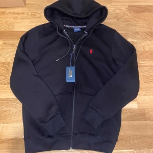 Svart hoodie från Polo Ralph Lauren - Svart hoodie från Polo Ralph Lauren med röd broderad logga på bröstet. Tröjan har dragkedja framtill, snörning i huvan och ribbade muddar. Klassisk och stilren design, perfekt för dig som gillar streetwear och premiumkänsla.
