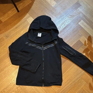 Svart vindjacka med huva från Nike - Svart vindjacka från Nike med hel dragkedja, huva och diskreta grå reflexdetaljer över bröstet. Jackan har en ficka med dragkedja på ena ärmen och är perfekt för dig som gillar sportig stil. Snygg och enkel design med Nike-logga på bröstet.