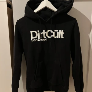 Svart Dirt Cult hoodie - Svart hoodie från Dirt Cult med stor vit logga och texten San Diego på bröstet. Klassisk modell med huva, snörning och känguruficka. 