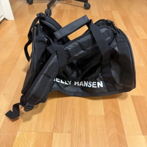 Svart Helly Hansen 30L duffelväska - Säljer en helt nytt oanvänd svart duffelväska från Helly Hansen på 30 liter. Väskan har tydlig HH-logga i vitt, bärhandtag och axelremmar så den kan bäras som ryggsäck. Tillverkad i slitstark syntet och perfekt för träning eller resor.
