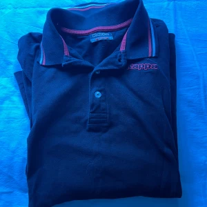 Svart pikétröja från Kappa M - Svart pikétröja från Kappa med röda och vita detaljer på krage och ärmslut. Klassisk pikékrage, knäppning med två knappar och broderad Kappa-logga på bröstet. Perfekt för en sportig och stilren look.