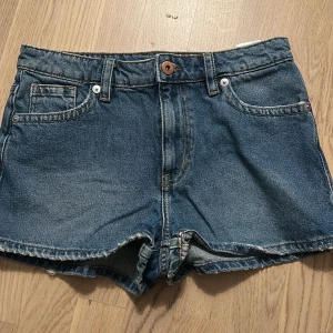 Klassiska blå jeansshorts - Snygga blå jeansshorts med fem fickor, bälteshällor och råa kanter nertill. Shortsen har klassisk passform och orange kontrastsömmar. Perfekta för varma dagar och enkel att matcha med allt.