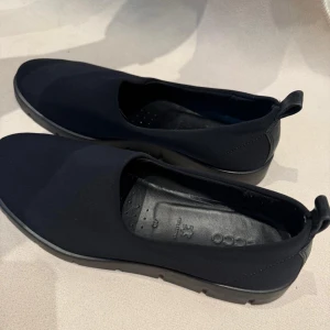 Svarta ballerinaskor från ECCO - Svarta ballerinaskor från ECCO med stilren design och rund tå. Ovandel i mjukt material och innersula i äkta skinn för extra komfort. Skorna har en platt sula med ECCO-logga undertill och en liten ögla bak för enkel påtagning.