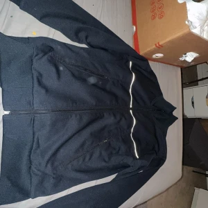 Svart Moncler jacka med dragkedja S - Svart jacka från Moncler i storlek S med hel dragkedja framtill, två snedställda fickor med dragkedja och en vit reflexdetalj över bröstet. Jackan har ståkrage och Moncler-logga på ärmen. Insidan har orange meshfoder för extra komfort.