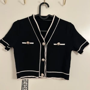 Svart kortärmad topp med vita detaljer från SHEIN  - Svart topp med korta ärmar och vita kontrastkanter i storlek XS från SHEIN. Toppen har dekorativa knappar framtill och två fejkfickor med knappar. Croppad modell med v-ringning. 
