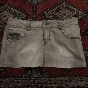 Grå jeanskjol med dragkedjor - Säljer en grå jeanskjol från Culte med rå kant nertill och coola dragkedjor på både fram- och bakfickor. Kjolen har klassisk femficksdesign, bälteshällor och en snygg, rak passform. Perfekt för dig som gillar en edgy streetstil.