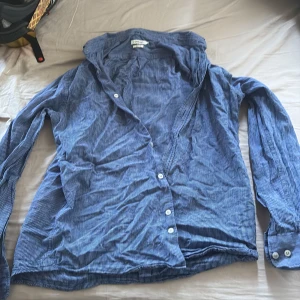 Blå randig skjorta från J.Lindeberg - Slim fit skjorta från J.Lindeberg i blått med tunna vita ränder. Skjortan har klassisk krage, långa ärmar med manschett och vita knappar framtill. Perfekt för dig som gillar en stilren och fräsch look.