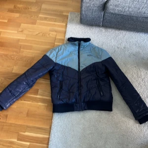 Blå pufferjacka med dragkedja oanvänd - Snygg pufferjacka från Everest i mörkblå och ljusblå färgblock. Jackan har hög krage, två fickor med dragkedja och quiltad design. Perfekt för kyliga dagar och har en modern look som passar till jeans och sneakers.