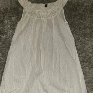 Vit kortklänning från Vero Moda - Supersöt vit kortklänning från Vero Moda med diskreta ränder och ett brett, virkat spetsmönster vid halsen. Klänningen är ärmlös och har en luftig passform, perfekt för varma dagar. Materialet känns lätt och bekvämt mot huden.