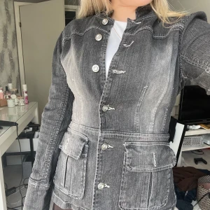 Svart/grå jeansjacka från Pastille Menthe - Snygg svart jeansjacka från Pastille Menthe med markerade sömmar och silverfärgade knappar. Jackan har två stora fickor framtill (som också har samma knappar med syns ej på bild), figurnära passform och lång ärm. Perfekt för dig som gillar en cool och stilren look.