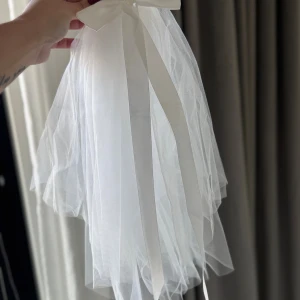 Vit diademslöja med rosett - Snygg vit slöja på diadem med stor rosett upptill och långa band. Slöjan är gjord i tunn tyll och fästs enkelt med metallkam. Perfekt för möhippa osv, ger en riktigt drömmig vibe till din look. Aldrig använd finns i Vasastan 