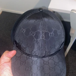 Svart Gucci keps med GG-mönster - Snygg svart keps från Gucci med klassiskt GG-mönster över hela framsidan. Kepsen har mesh på baksidan och justerbar snapback. Materialet är polyester och har en lätt glansig finish som ger en lyxig vibe. Perfekt för dig som vill ha en statement-accessoar.