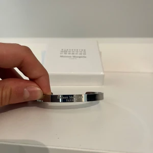 Maison Margiela silverfärgat armband - Stilrent armband från Maison Margiela i silverfärgad metall med graverad logotyp på framsidan. Armbandet har en minimalistisk och modern design, perfekt för dig som gillar diskreta men exklusiva accessoarer. Levereras i original ask.