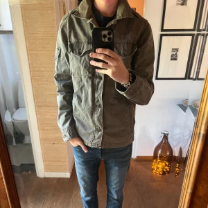 Olivgrön fieldjacka från Hilfiger Denim - Snygg fieldjacka från Hilfiger Denim med klassisk krage, axelklaffar och fyra fickor framtill. Jackan har dragkedja och knappar framtill.Perfekt för dig som gillar en avslappnad och stilren look. Size medium.  Bra skick endast att en av knapparna på fick har gått av och en lapp inuti har klippts av, se bild. Det är även väldigt lite vit färg på ena ärmen vilket knappt syns bara små fläckar. Går antagligen att tvätta bort lätt.Bara att skriva vid frågor!😃