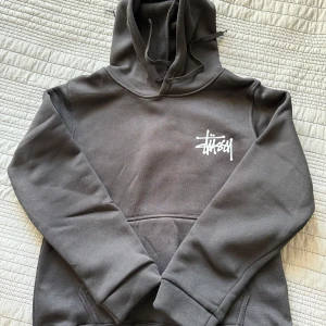Svart Stüssy hoodie med logga - Säljer en svart hoodie från Stüssy med stor vit logga på ryggen och mindre logga på bröstet. Tröjan har huva med snörning och en klassisk känguruficka framtill. Perfekt för dig som gillar streetwear och vill ha en stilren look.