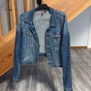 Blå jeansjacka från H&M Divided - Snygg klassisk blå jeansjacka från H&M Divided i storlek 42. Jackan har långa ärmar, knappar framtill och två bröstfickor med knapp. Den har en cool, lite tvättad look och är perfekt för att ge din outfit en avslappnad vibe.