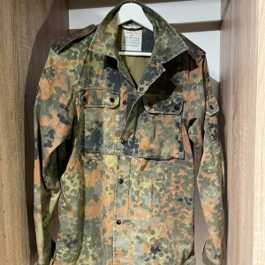 Camouflage overshirt - Säljer en grön, brun och svart camouflage overshirt med klassisk krage och bröstfickor med knappar. Skjortan har långa ärmar och axelklaffar. Materialet är slitstark bomull. Pris satt ex frakt
