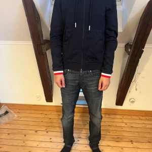 Mörkblå hoodiejacka från Moncler - Snygg mörkblå hoodiejacka från Moncler med dragkedja, huva och snörning. Jackan har två fickor fram och Moncler-logga på vänster ärm. Muddarna har coola rödvita detaljer som sticker ut. Perfekt för dig som gillar stilren streetwear.
