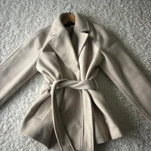 Beige kappa med bälte från Vero Moda - Stilren beige kappa från Vero Moda med klassisk krage och knytskärp i midjan. Jackan har långa ärmar och är tillverkad i ett mjukt ullblandat material. Perfekt för dig som gillar en clean och tidlös look.