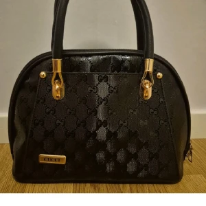 Svart handväska  med guldiga detaljer - Snygg svart handväska från Cucci med klassiskt monogrammönster och guldiga metalldetaljer. Väskan har två handtag, dragkedja upptill och rymligt fack inuti. Materialet är glansig syntet med struktur och väskan har en elegant, rundad form.