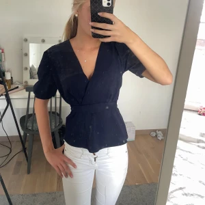 Mörkblå omlottblus med bälte - Snygg mörkblå omlottblus med korta puffärmar och markerad midja tack vare ett brett bälte. Blusen har v-ringning och en stilren, feminin siluett. Perfekt att styla med jeans eller kjol för en trendig look.