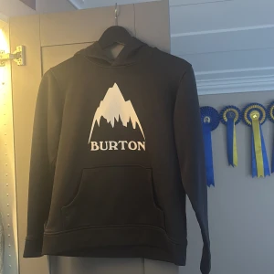 Svart Burton hoodie med vit logga - Svart hoodie från Burton i storlek M med stor vit logga och bergmotiv på bröstet. Tröjan har huva, magficka och långa ärmar. Perfekt för dig som gillar streetwear och vill ha en chill look. Materialet är mjukt och känns skönt mot huden.