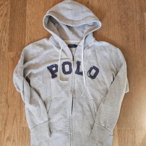 Grå hoodie från Polo Ralph Lauren - Snygg grå hoodie från Polo Ralph Lauren med dragkedja och stor huva. Tröjan har vita snören och mörkblåa bokstäver 'POLO' i tyg framtill. Perfekt för chill dagar och enkel att matcha med jeans eller joggers.